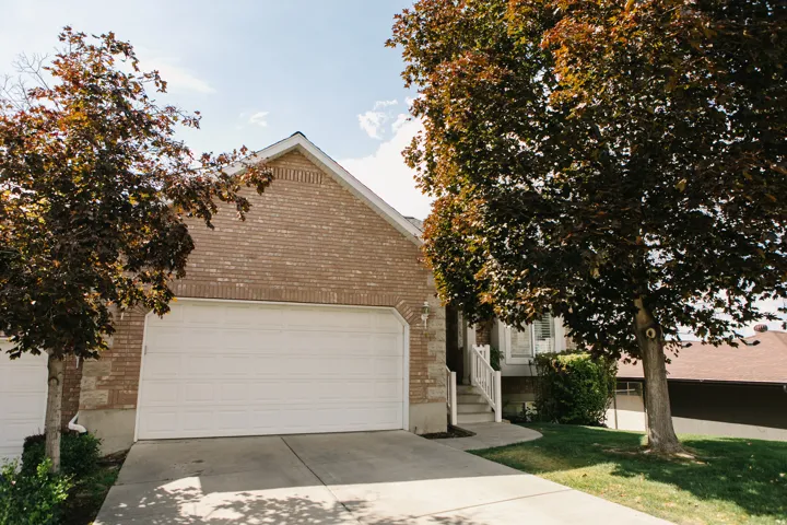 Brick home featuring a two-car garage, concrete driveway, front entry stairs with white railings, and mature trees providing natural landscaping