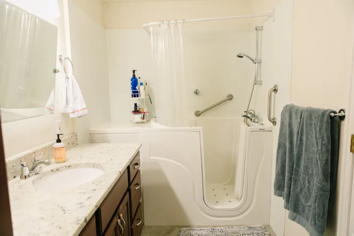 Bathroom featuring a walk-in tub with grab bars, a shower wand, a vanity with a white countertop and integrated sink, and dark wood cabinetry with silver hardware