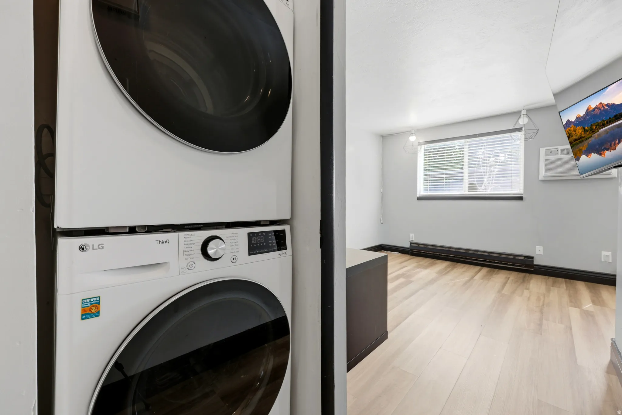 Stackable washer and dryer unit, light wood-finish flooring, recessed window with blinds, baseboard heating, and an installed wall-mounted air conditioning unit