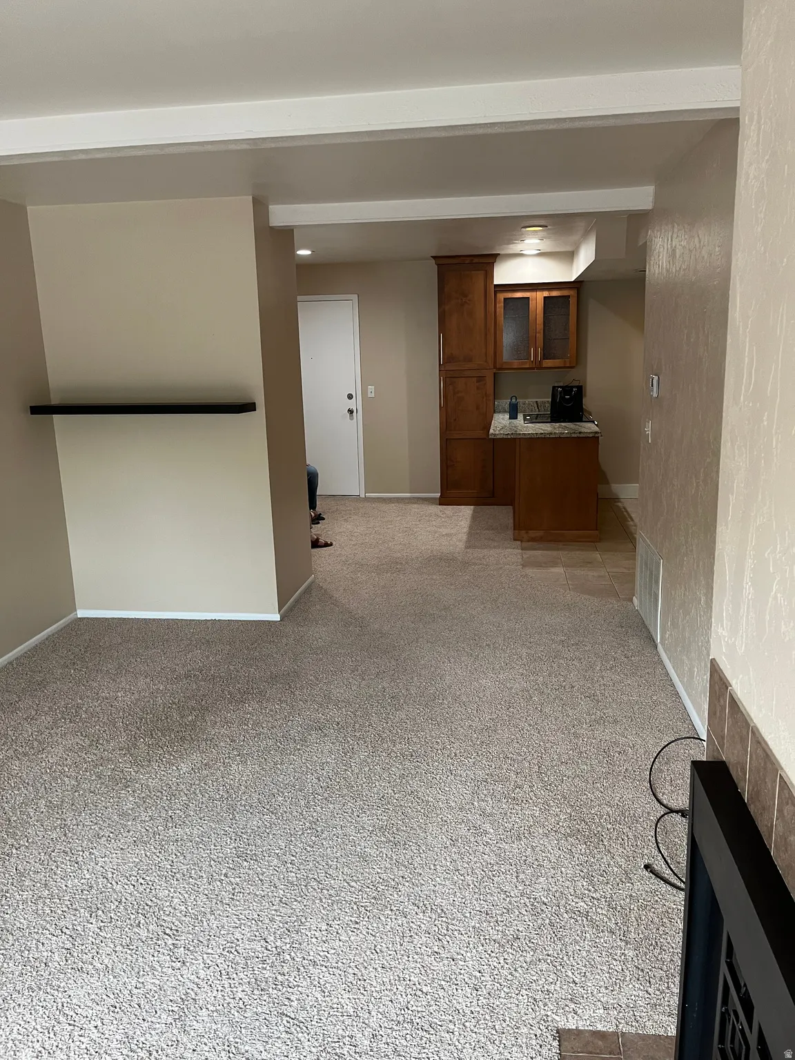 Neutral-toned carpeting, recessed lighting, wood-finish cabinetry with glass-front accents, tiled flooring in the kitchen area, and a tiled fireplace surround