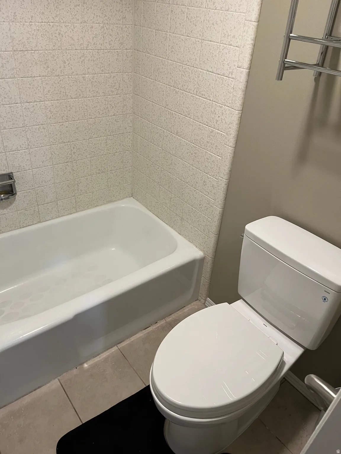 White bathtub with textured wall tiling, a modern toilet, and light-toned floor tiles
