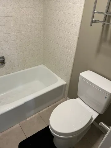 White bathtub with textured wall tiling, a modern toilet, and light-toned floor tiles