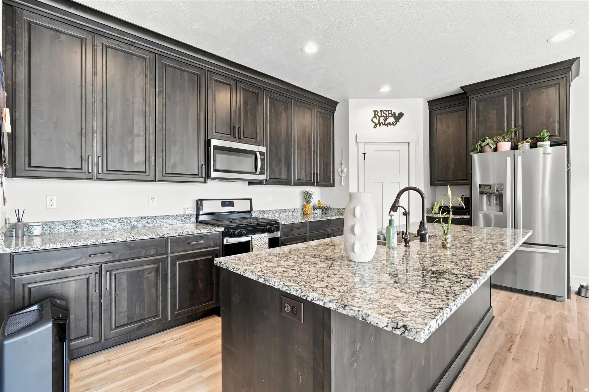 Spacious kitchen featuring dark wood-finish cabinetry, light speckled countertops, stainless steel appliances, a center island with a sink, and light wood-finish flooring
