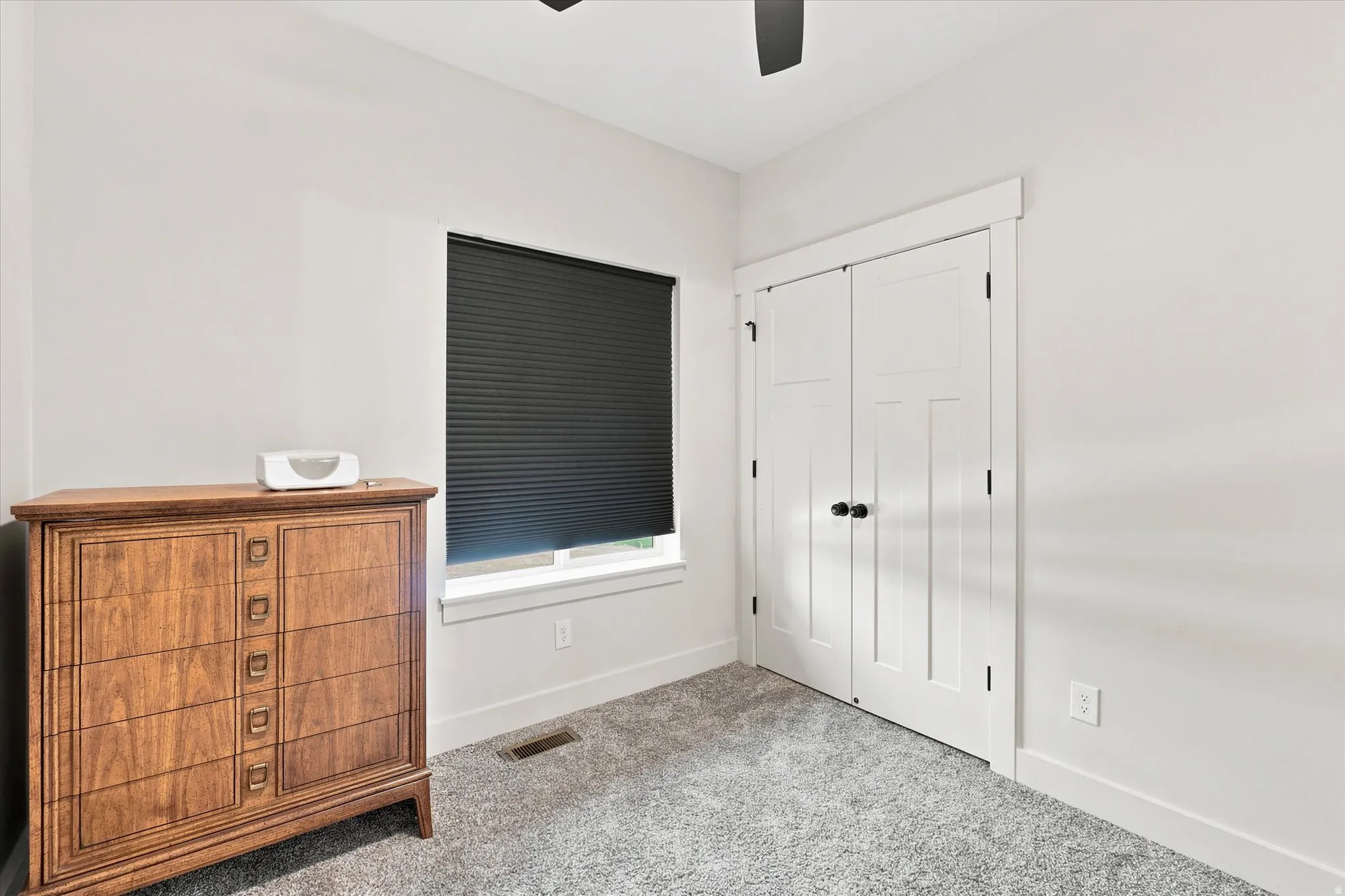 Light gray wall paint, gray carpet, white trim, window with dark cellular shades, and a two-panel closet door with matte black hardware