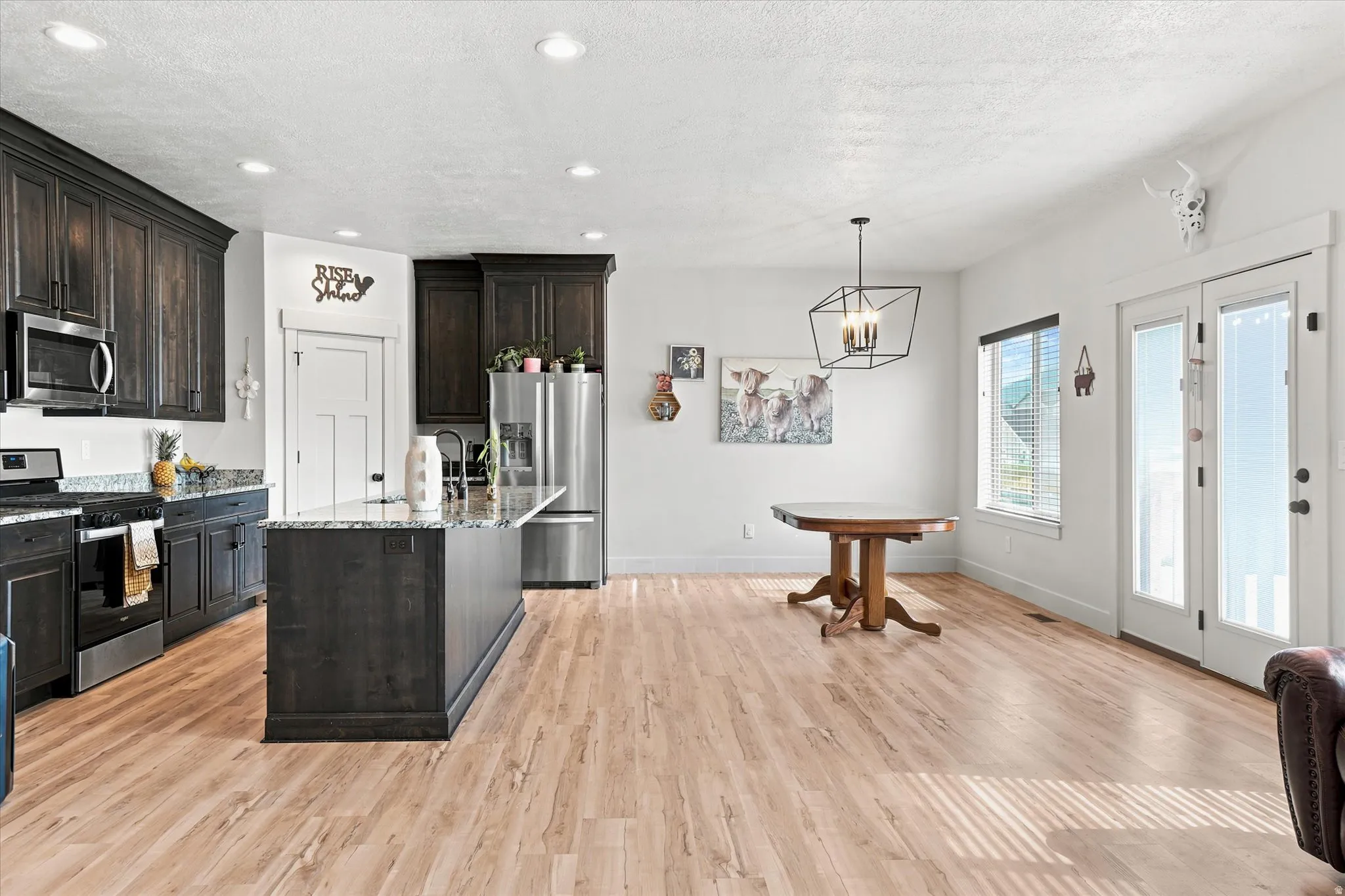Open-concept kitchen and dining area featuring dark wood cabinetry, stainless steel appliances, granite countertops, and light wood-finish flooring