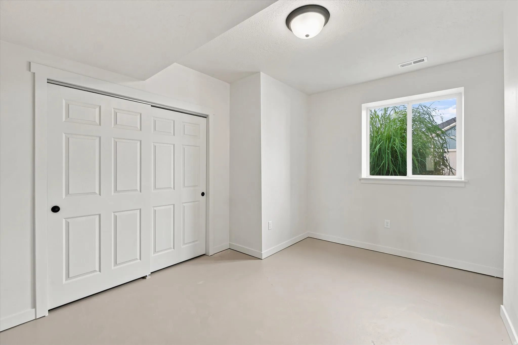 Bright interior space featuring a window with exterior foliage, neutral-toned flooring, white walls, and a ceiling-mounted light fixture
