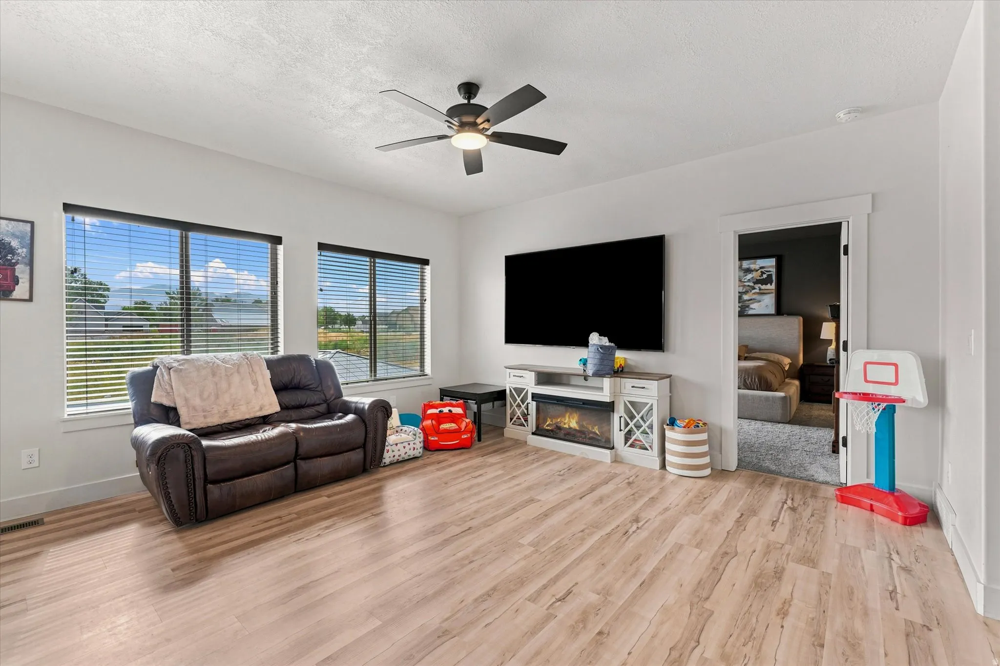 Spacious room featuring light wood-finish flooring, multiple windows with blinds, a built-in electric fireplace media console, and a ceiling fan with integrated lighting
