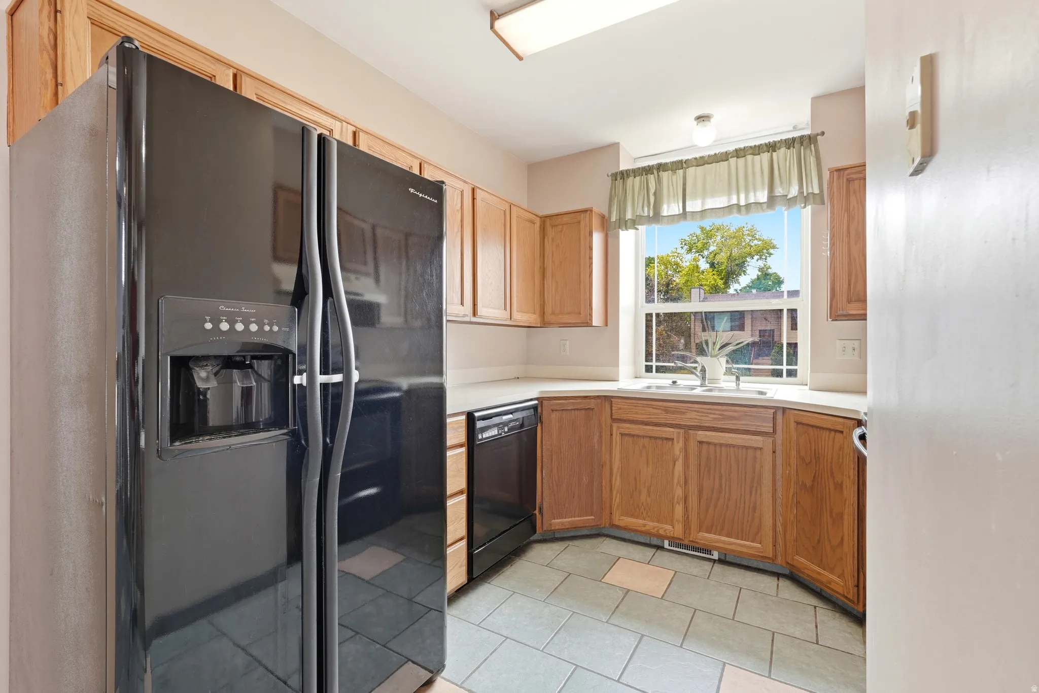 Kitchen featuring a black side-by-side refrigerator, warm wood-finish cabinetry, a corner sink with a double window, light-colored countertops, and tiled flooring