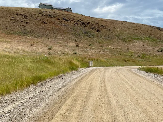 Gravel road ascending a natural incline, with a residential structure visible on the distant hillside