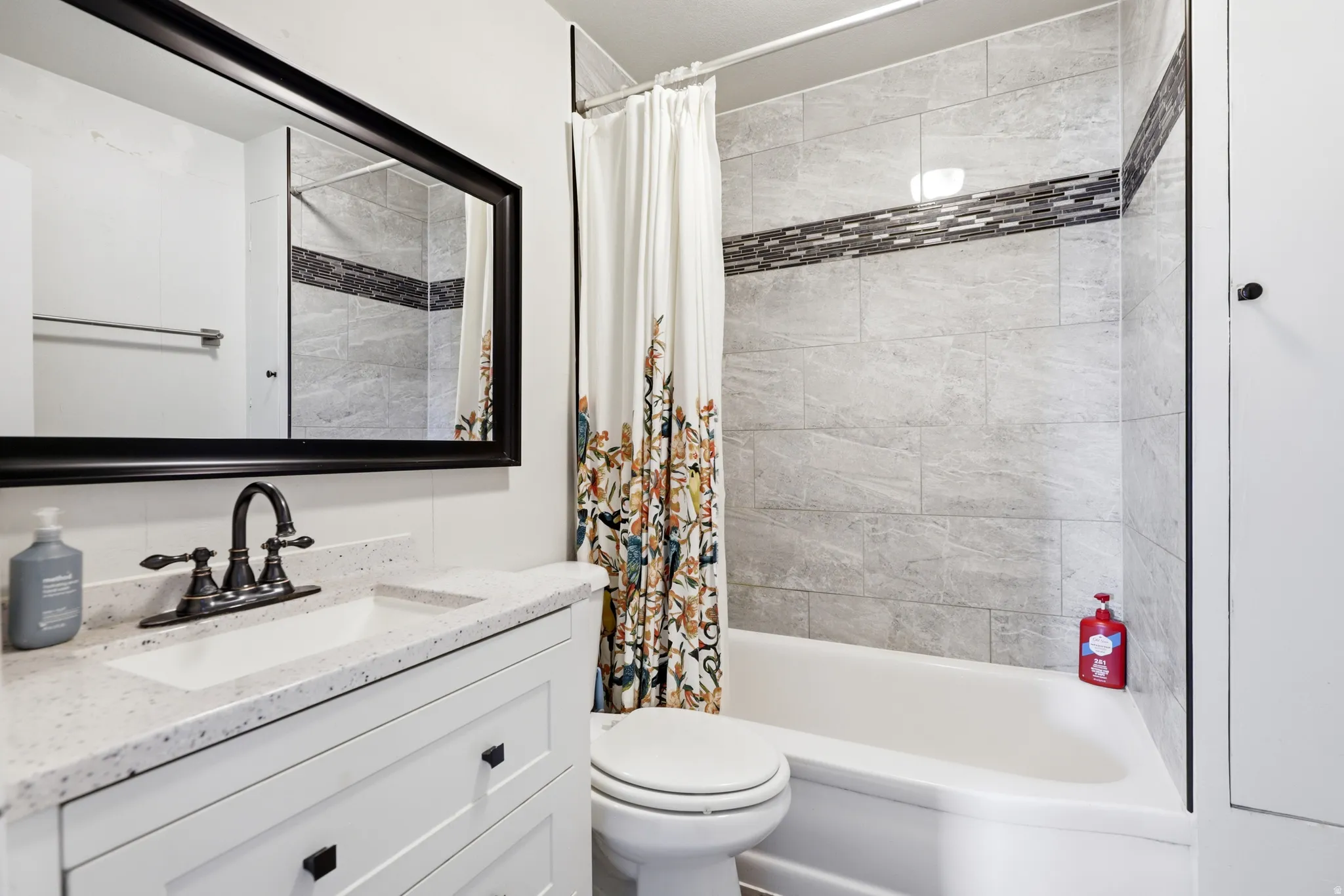 Contemporary bathroom featuring a white vanity with dark hardware, a spacious countertop, a dark-framed mirror, and a tub-shower combination with light-toned wall tiling and a decorative mosaic accent band