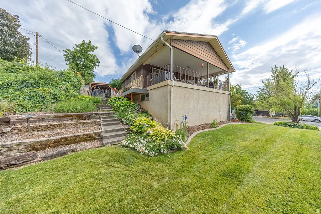 Exterior featuring a covered upper-level balcony, a lower-level patio area, a terraced garden with stone retaining walls, concrete stairs, and a lush green lawn