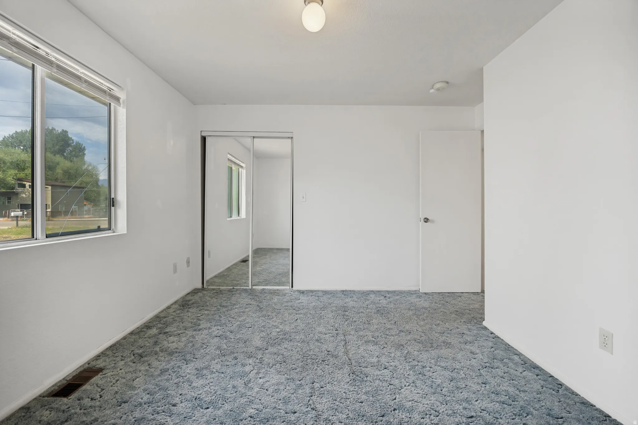 Spacious room featuring a large window, light blue carpet, white walls, and a mirrored closet door