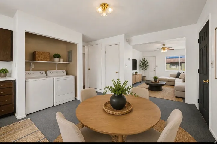 Integrated laundry area with shelving, overhead ceiling light fixture, kitchen cabinetry with white countertop, dining area with round wood table, and living area with window