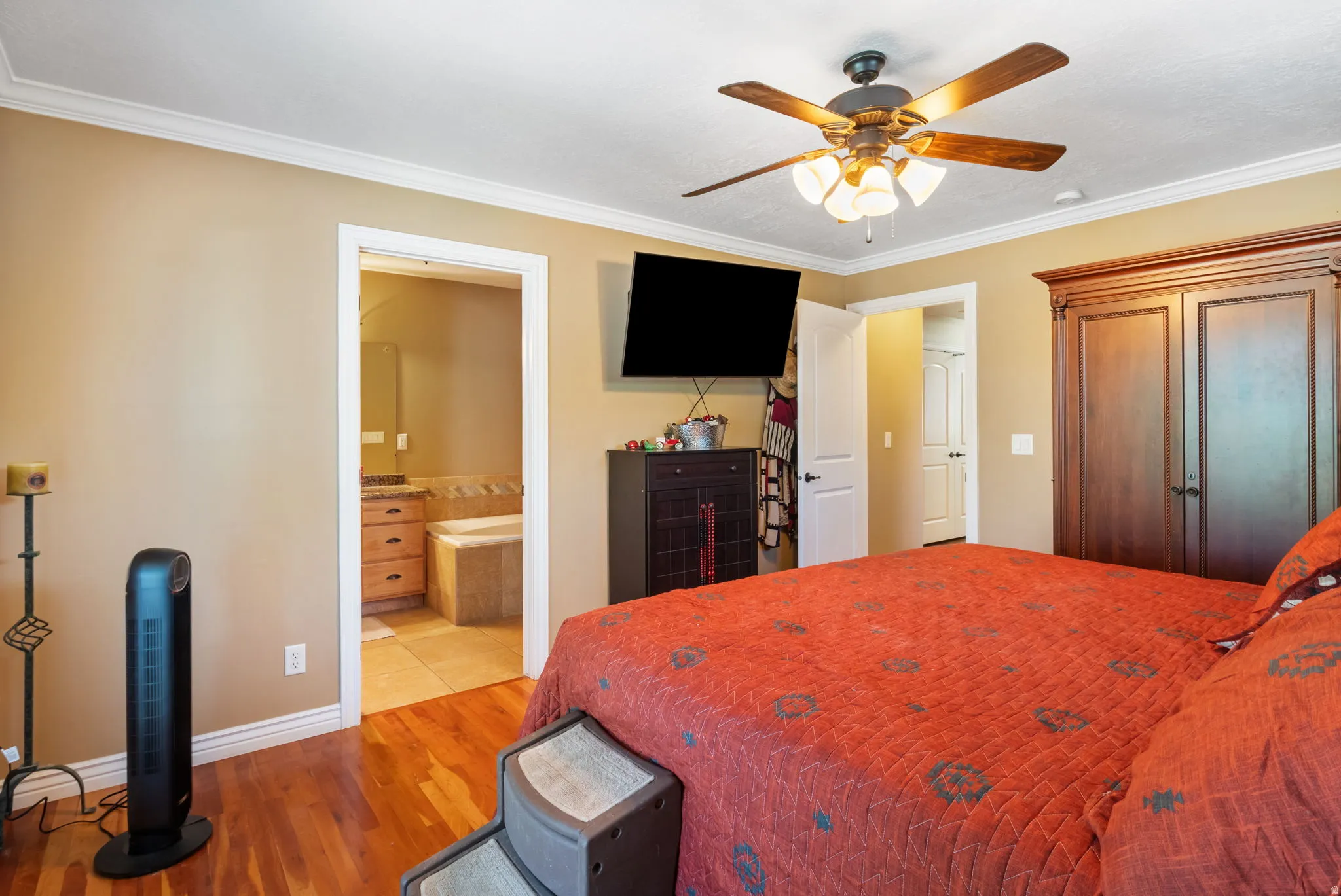 Spacious bedroom featuring wood-finish flooring, crown molding, a ceiling fan with integrated lighting, and an attached bathroom with a built-in tub and vanity