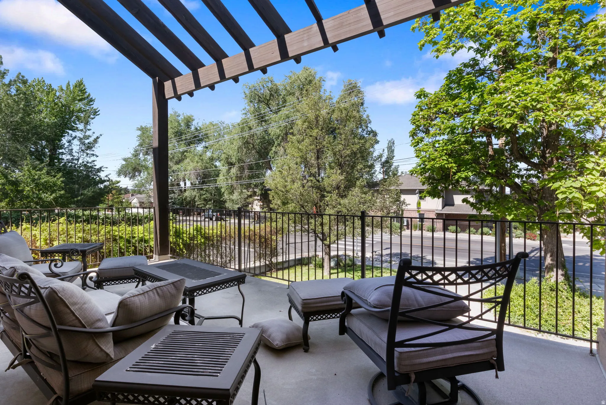 Spacious outdoor patio featuring a slatted pergola, concrete flooring, and black metal railings, surrounded by mature trees
