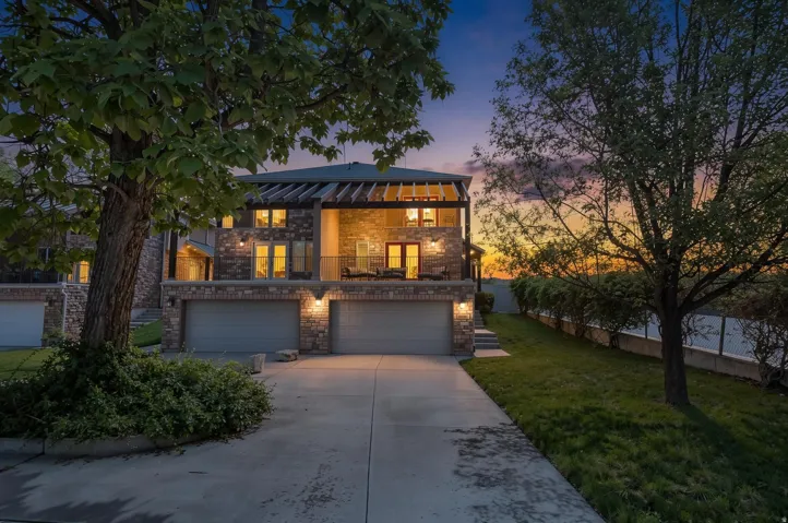 Multi-level residence featuring a stone and brick facade, multiple garage bays, an upper-level balcony with a pergola, exterior lighting, and a paved driveway