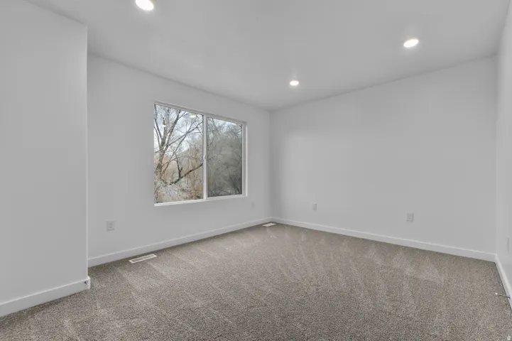 Spacious room featuring neutral carpeting, white walls, recessed ceiling lighting, and a prominent window with a natural backdrop