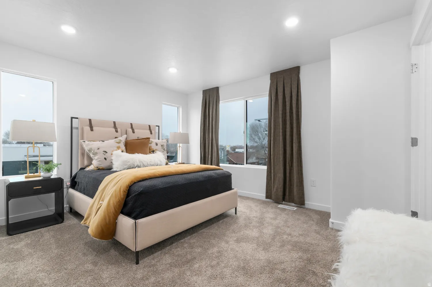 Carpeted bedroom featuring recessed lighting, a modern bed frame with a light-colored headboard, and multiple windows with full-length drapes