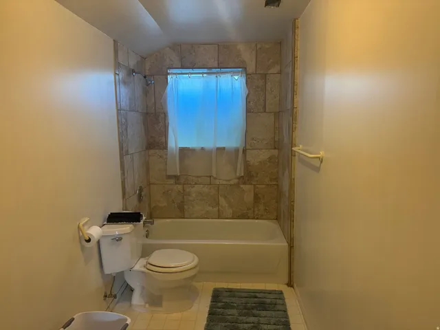 Bathroom featuring a white bathtub with a tiled surround, a white toilet, and a window with a sheer curtain