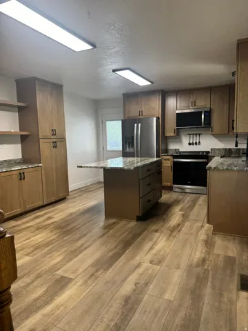 Kitchen featuring an island with ample storage, stainless steel appliances, wood-finish flooring, natural wood cabinetry, and granite-style countertops