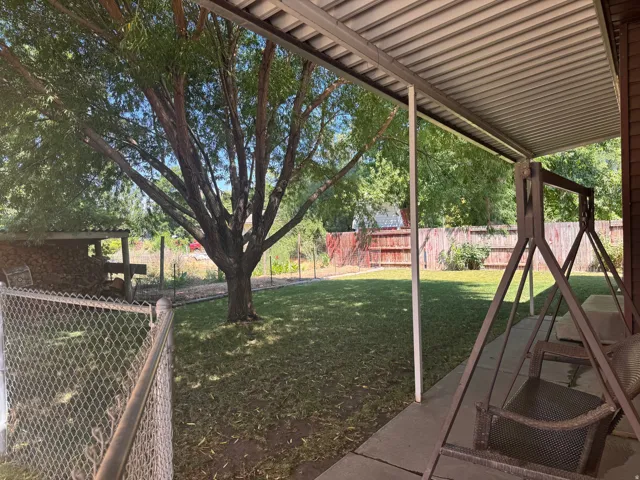 Covered patio area with a concrete slab, expansive lawn, mature shade tree, chain-link fence, and a wood privacy fence bordering the property