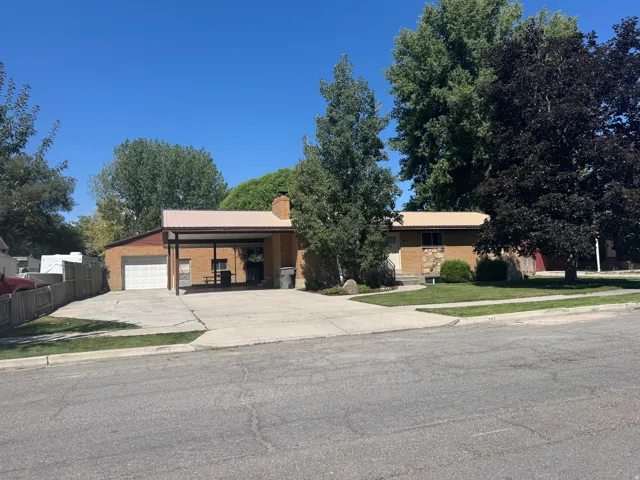 Brick exterior residence featuring a carport with a corrugated metal roof, an attached garage, a paved driveway, and mature landscaping