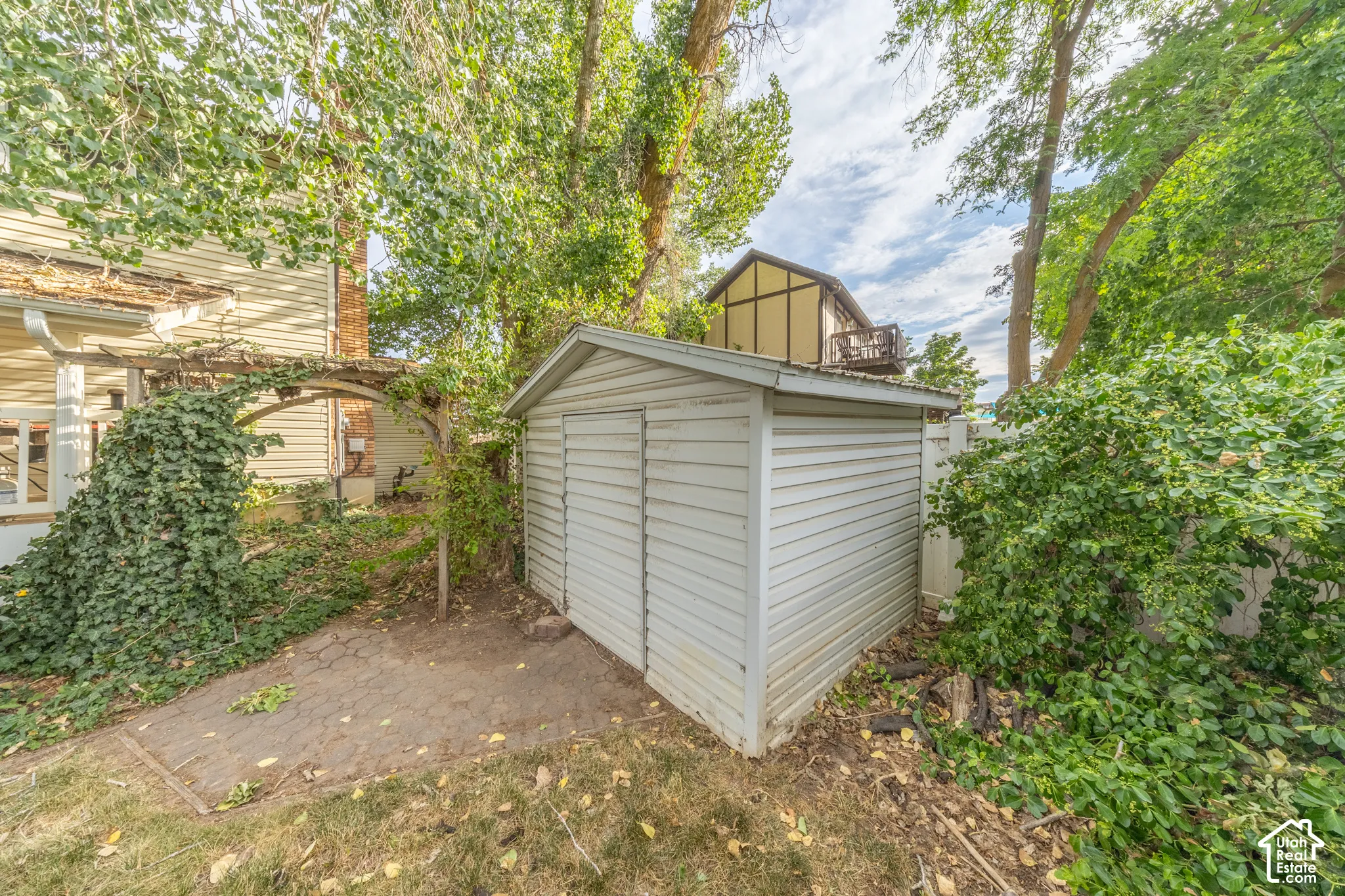 White sided shed with double doors, surrounded by mature trees, a privacy fence, and a paved path