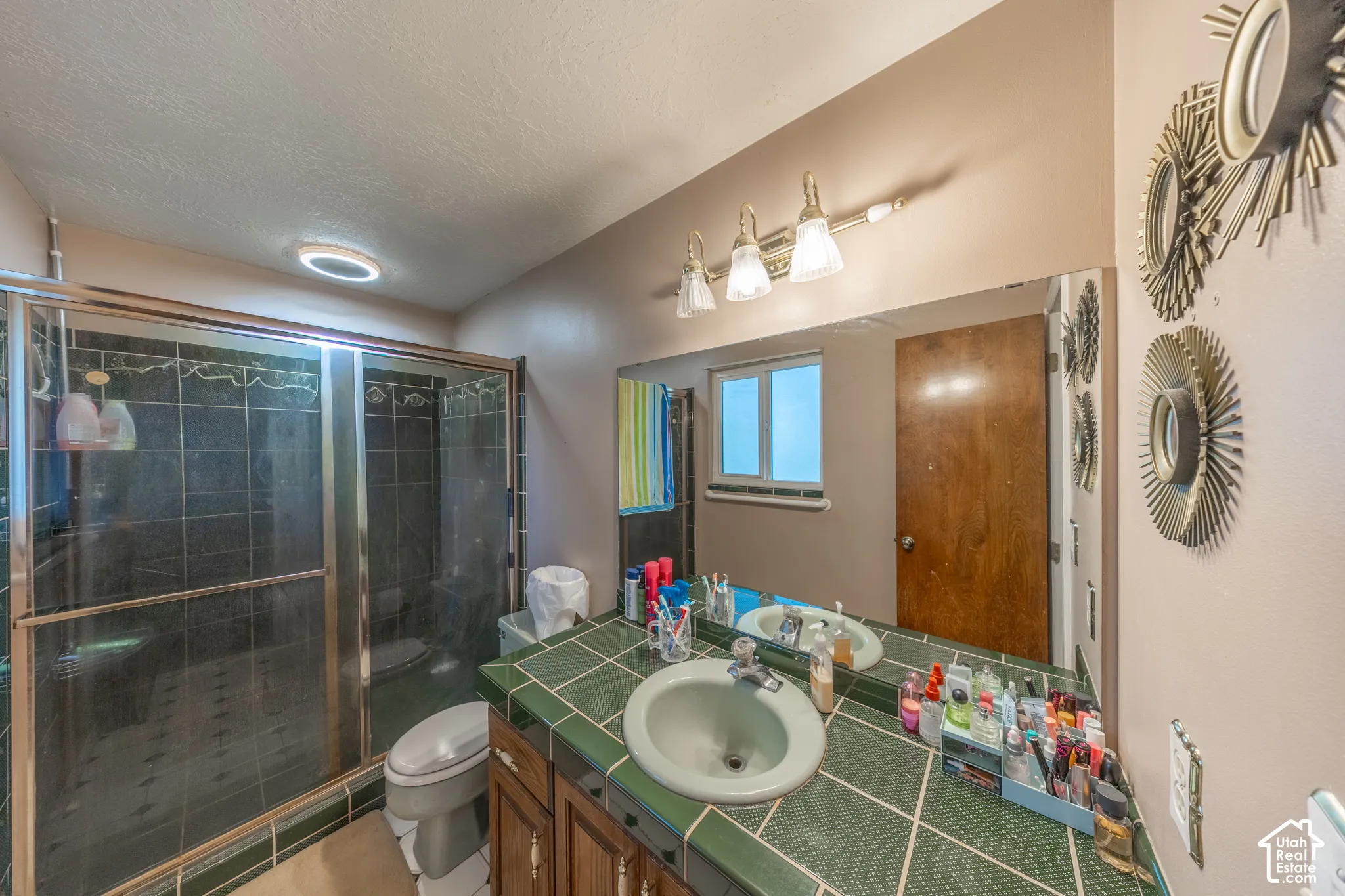 Glass-enclosed shower with dark tile surround, vanity with green tile countertop, integrated sink, wood-finish cabinetry, and wall-mounted vanity lighting