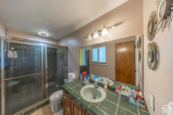 Glass-enclosed shower with dark tile surround, vanity with green tile countertop, integrated sink, wood-finish cabinetry, and wall-mounted vanity lighting