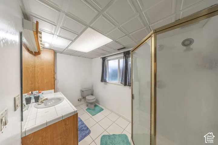 Bathroom featuring a tiled vanity with integrated sink, wood-finish cabinetry, a shower enclosure with gold-tone framing, white floor tiling, and a window