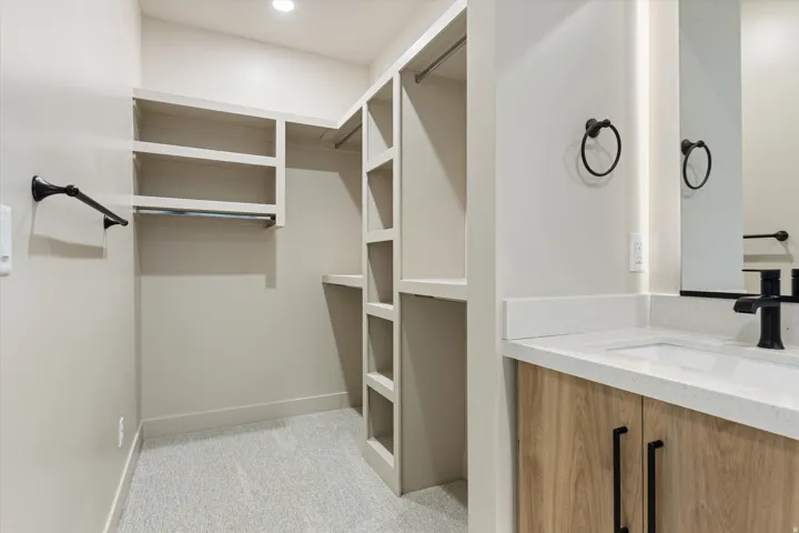 Walk-in closet and vanity area featuring custom built-in shelving, hanging rods, light-toned carpeting, a wood-finish vanity with a light-colored countertop, and a built-in mirror