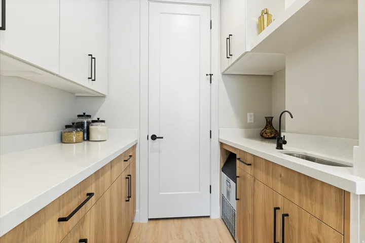 Pantry featuring light wood-finish lower cabinetry, white upper cabinets, light quartz countertops, and a stainless steel sink with a matte black faucet