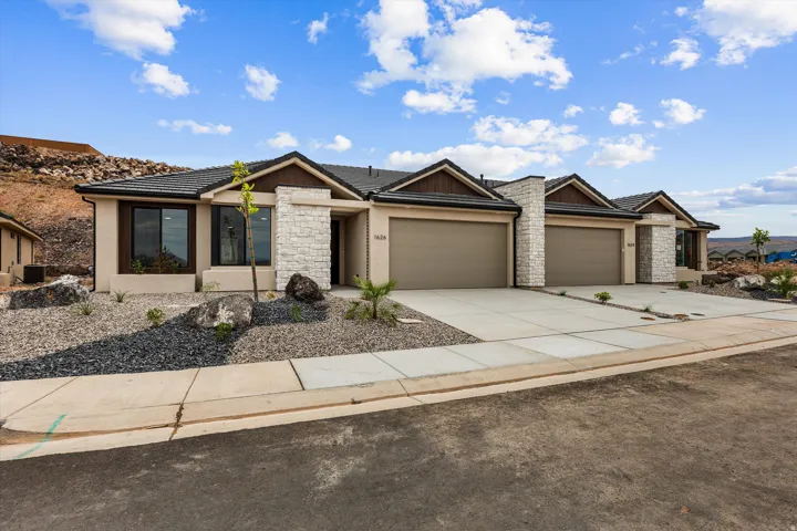 Contemporary residential exterior featuring stone veneer accents, stucco siding, a dark tile roof, two integrated garage doors, and a xeriscaped front yard with decorative rock and low-maintenance plantings