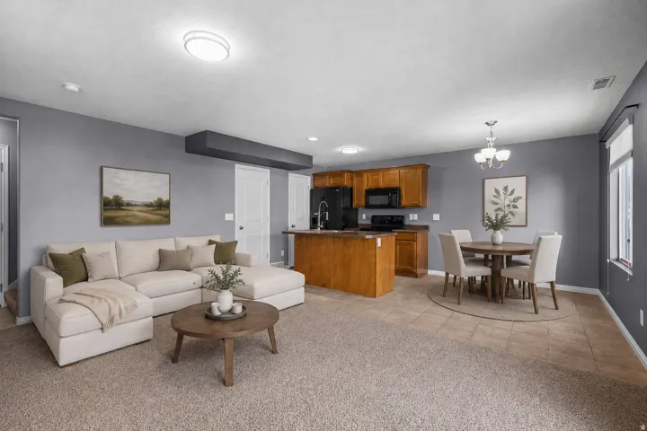 Open-concept living area featuring carpeted flooring, a kitchen island with seating, wood cabinetry, black appliances, and a window with blinds.  AI staged photo.
