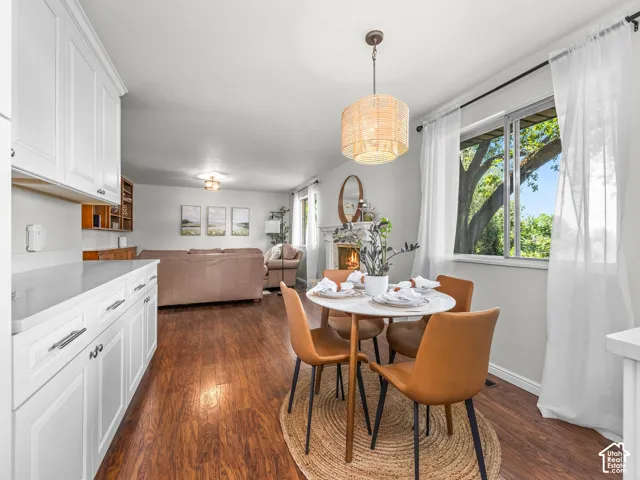 Open-concept living area featuring wood-finish flooring, white cabinetry with silver hardware, a dining area with a round table, a window with natural light, and a fireplace with a mantel