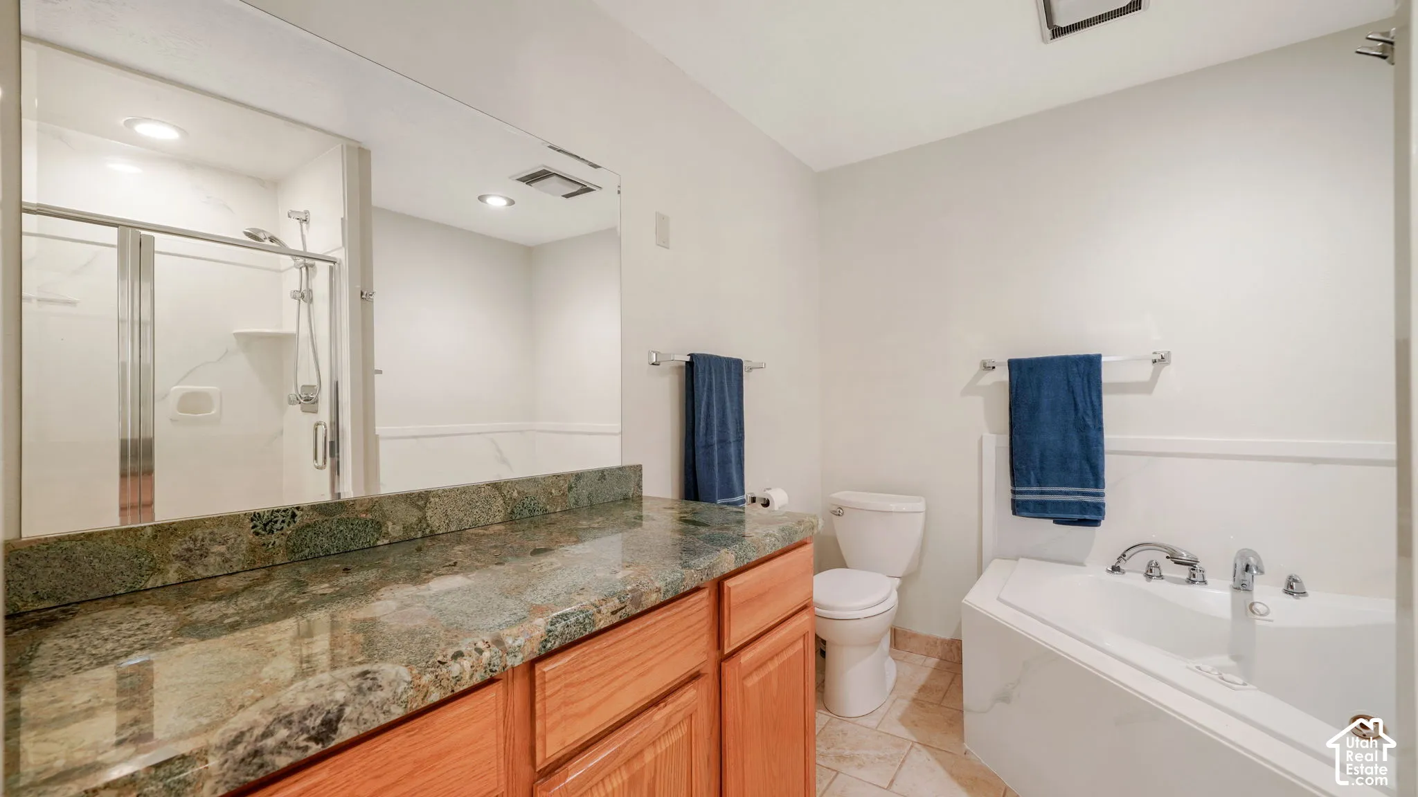 Bathroom featuring a corner soaking tub, glass-enclosed shower, wood-finish vanity with stone countertop, and tile flooring