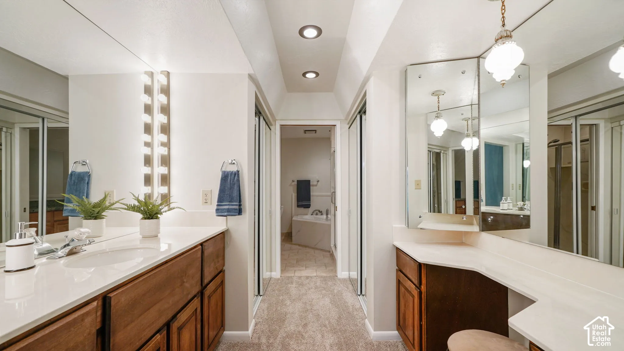 Spacious bathroom featuring dual wood-finish vanities with solid surface countertops, extensive mirror coverage, recessed ceiling lighting, and a separate vanity area with a decorative pendant light