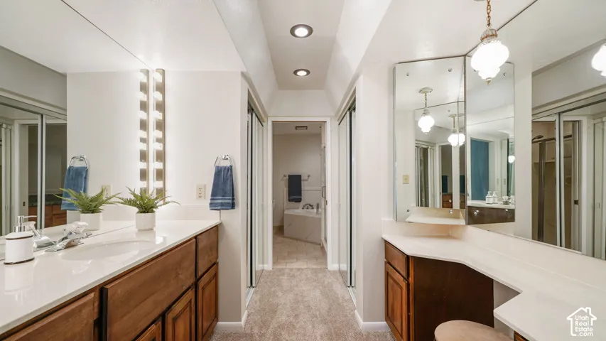 Spacious bathroom featuring dual wood-finish vanities with solid surface countertops, extensive mirror coverage, recessed ceiling lighting, and a separate vanity area with a decorative pendant light