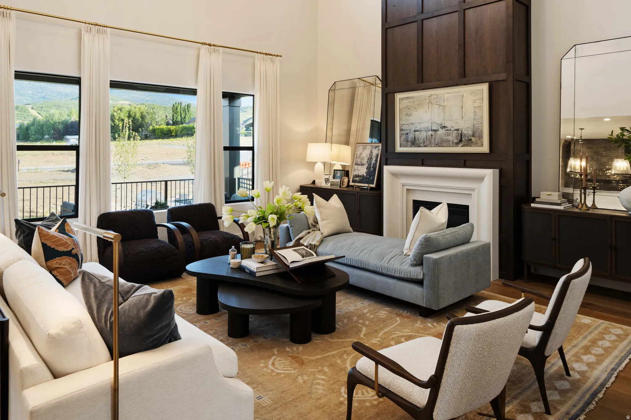 Spacious living area featuring a fireplace with a detailed mantel, dark wood paneling, large windows with black frames, and light-toned wood-finish flooring