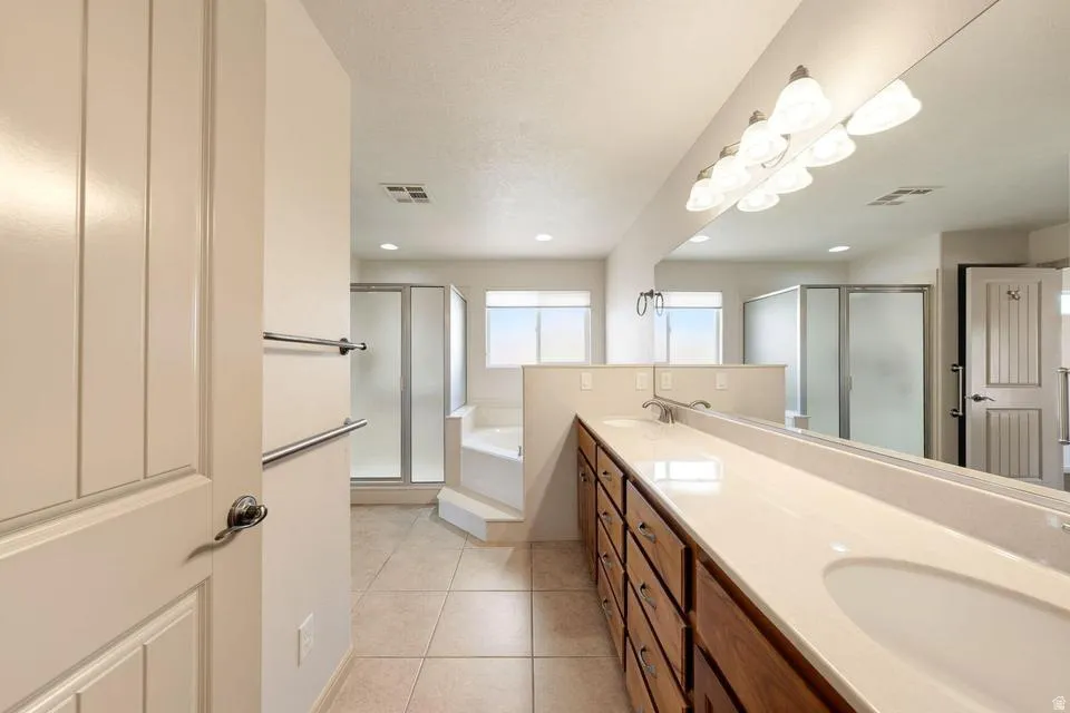 Bathroom featuring dual sinks set in a light-toned countertop, a large vanity mirror, tile flooring, and a walk-in shower with a glass enclosure