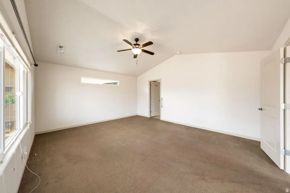 Spacious room featuring vaulted ceilings, a ceiling fan with integrated lighting, brown carpeting, multiple windows, and white interior doors