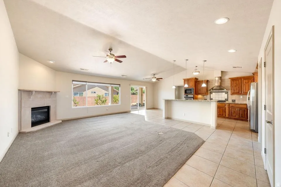 Vaulted ceiling great room featuring a corner fireplace, large window, and carpet flooring, opening to a kitchen with wood cabinetry, stainless steel appliances, and a breakfast bar