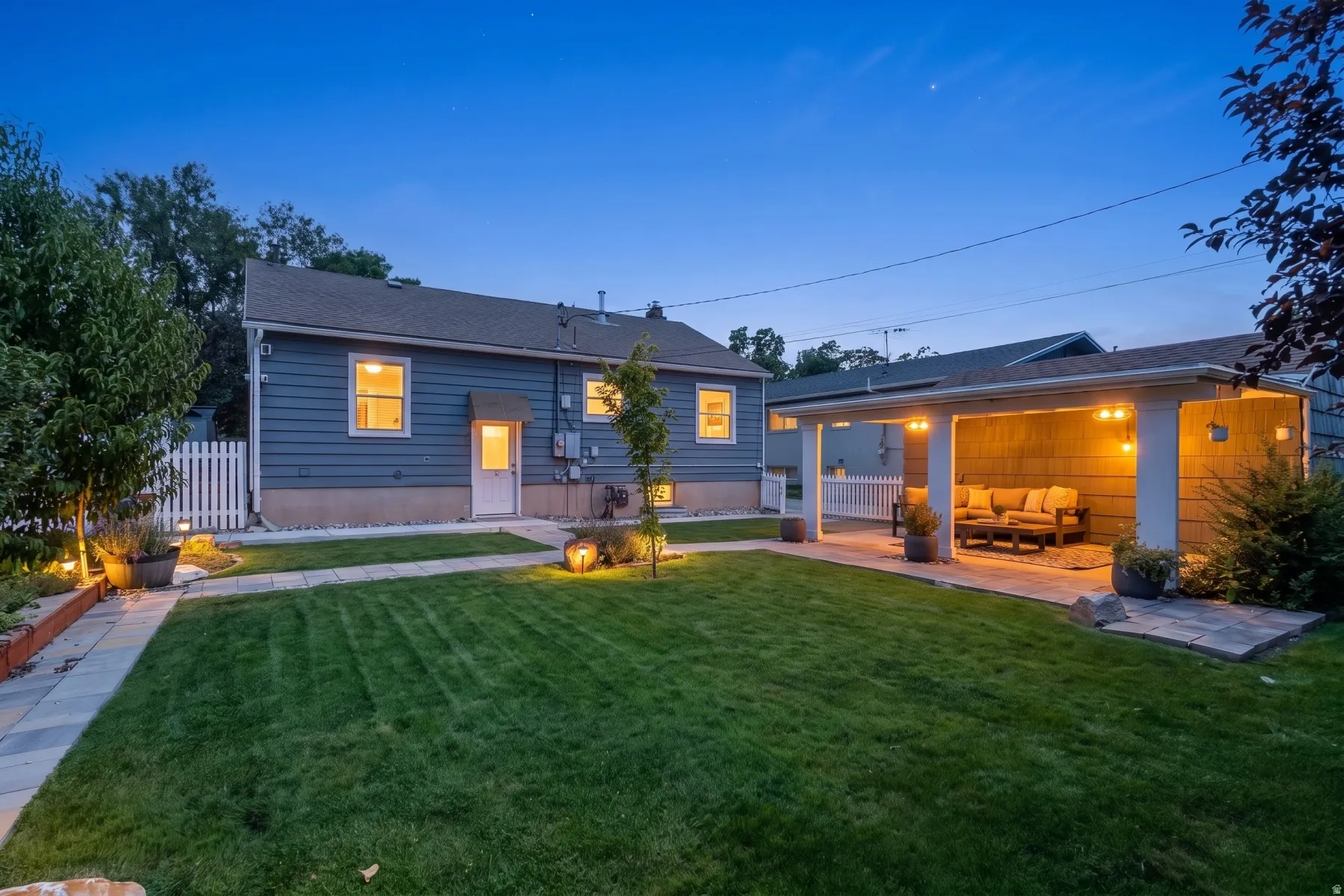 Expansive backyard featuring a covered patio with integrated lighting, a stone paver patio, mature landscaping, and a white picket fence