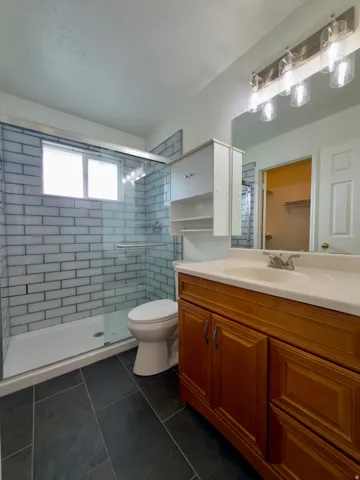 Primary bathroom featuring a large glass-enclosed shower with white subway tile, a vanity with a light-colored countertop and integrated sink, a wood-finish cabinet, and dark rectangular floor tiles