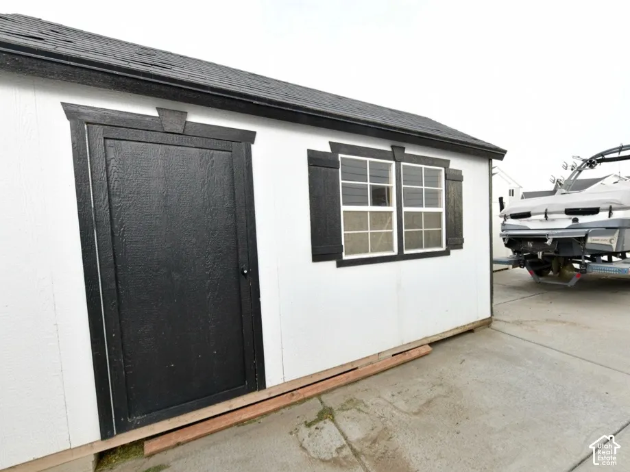 Detached storage shed featuring a black shingle roof, white siding, and a black paneled door