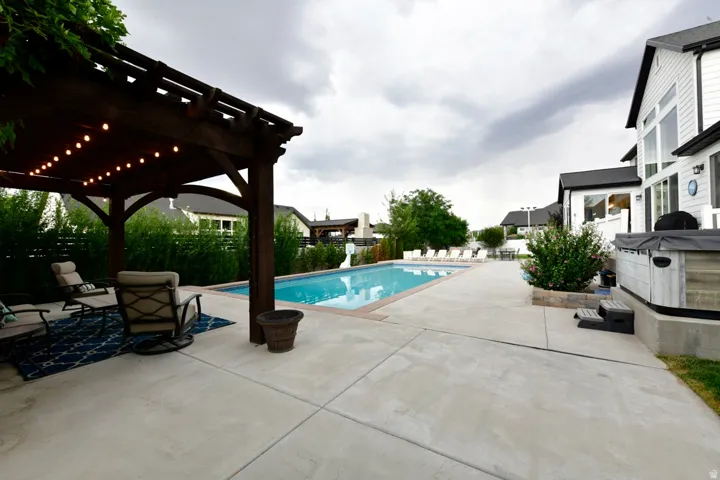 Expansive concrete patio featuring a rectangular swimming pool, a freestanding hot tub, and an attached pergola with string lighting