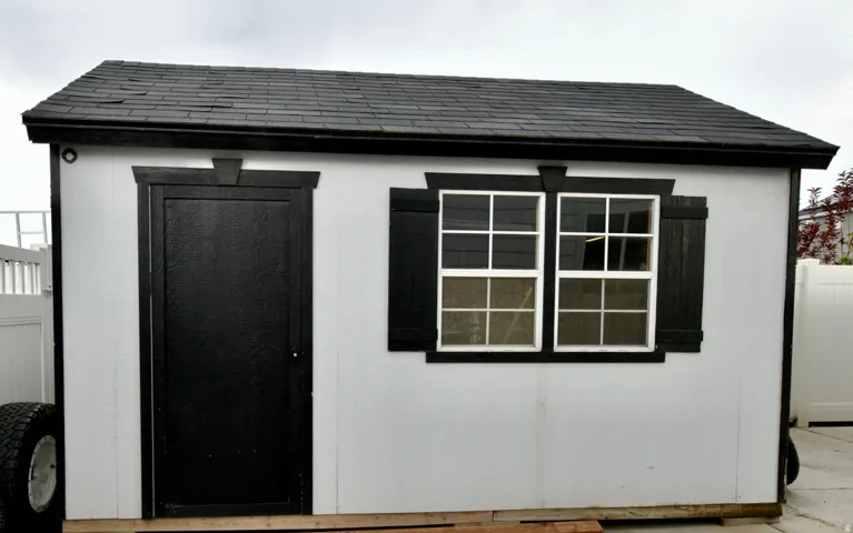 Detached shed featuring a black shingle roof, white siding, black trim, a black entry door, and two multi-pane windows with black shutters