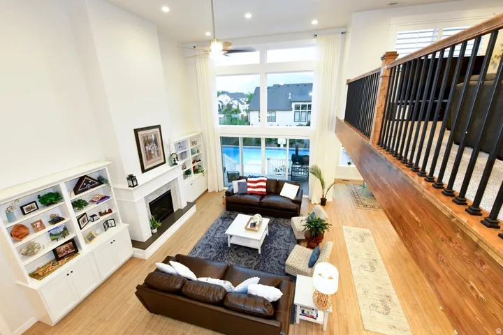 Two-story living room featuring a fireplace with white mantel, built-in shelving, wood-finish flooring, and a large window wall overlooking an outdoor pool