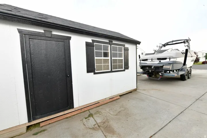 Detached storage shed featuring a black shingle roof, white siding, and a black paneled door