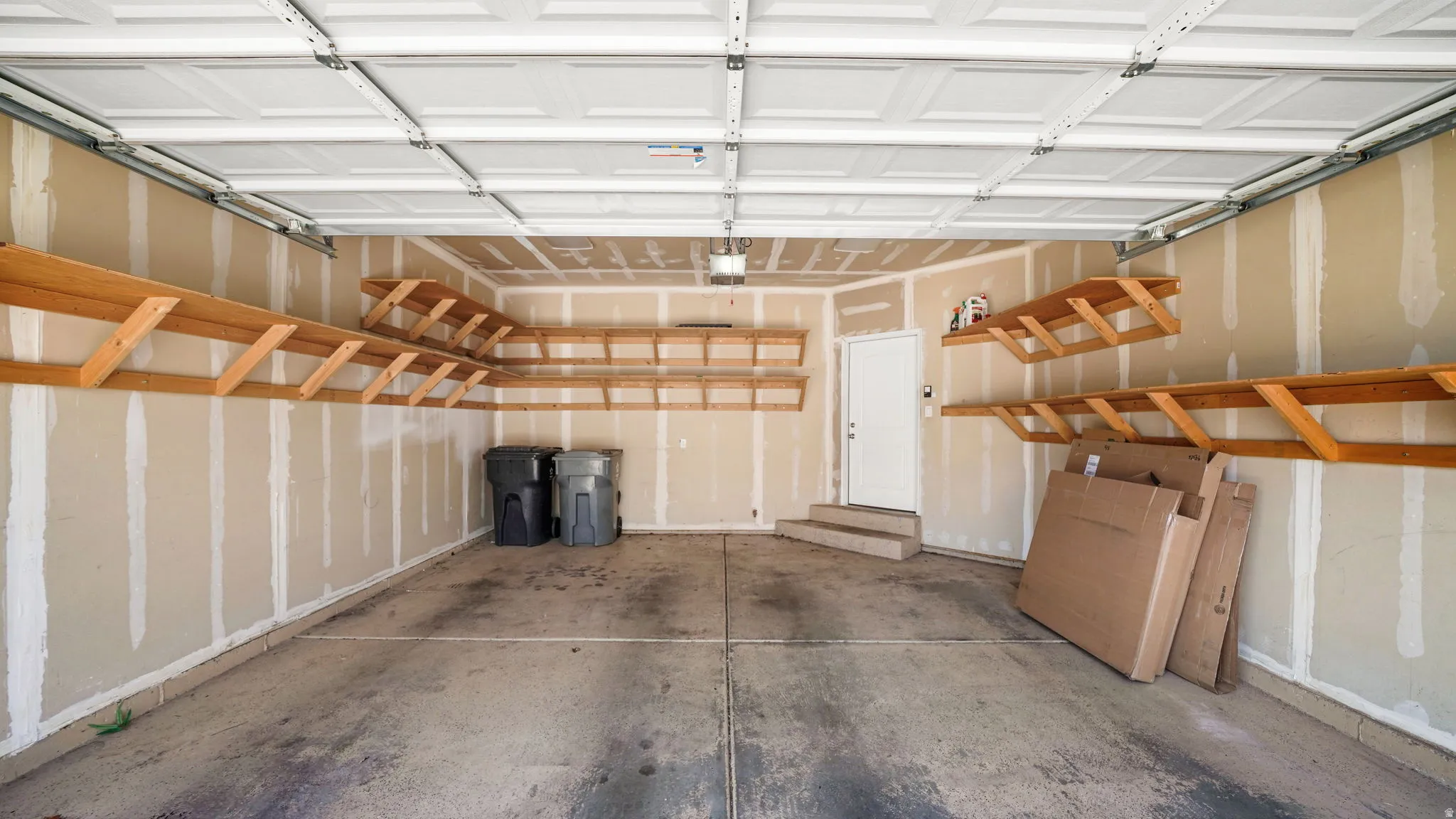 Spacious garage with concrete flooring, overhead garage door, built-in wooden shelving, and a white interior door with steps